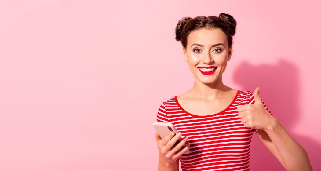 Junge Frau zeigt lachend "Daumen nach oben" in der anderen Hand hält sie ein Smartphone.